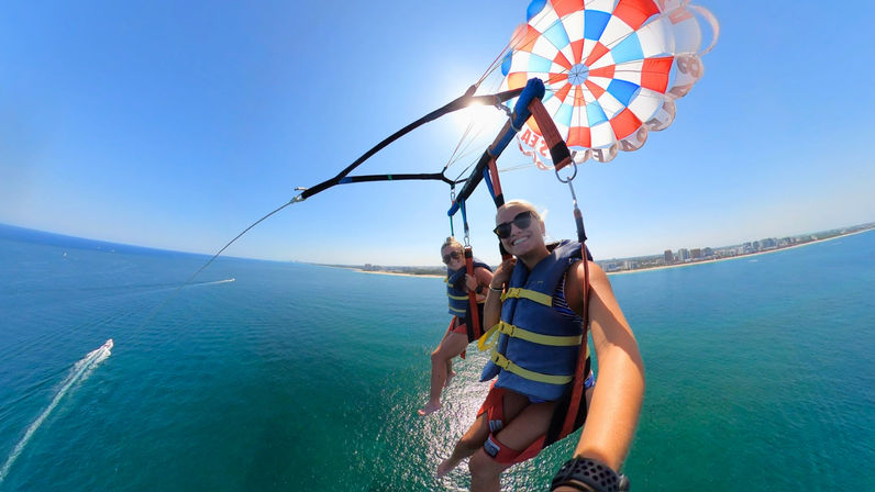 Two smiling parasail riders high above turquoise ocean, tethered to a red, white and blue parachute with a sandy coastline and beachfront skyline below on a sunny day