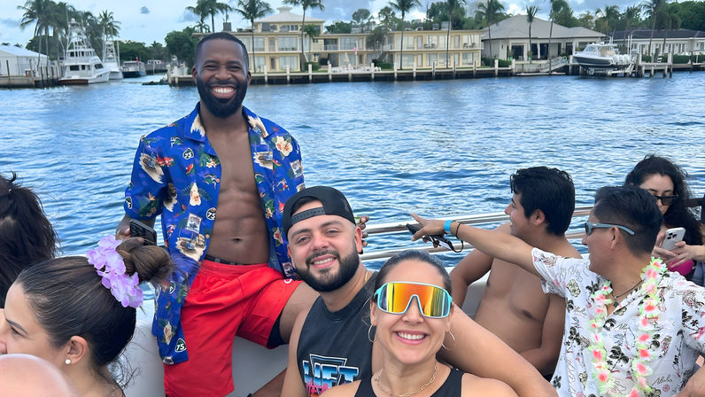 Smiling friends on a colorful boat party at a sunny waterfront marina with palm trees, yachts, and waterfront homes
