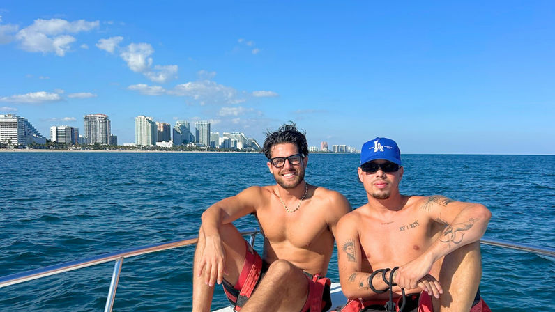 Two shirtless men relaxing on a boat in clear blue ocean with a coastal city skyline and sunny sky, summer boat vacation vibe