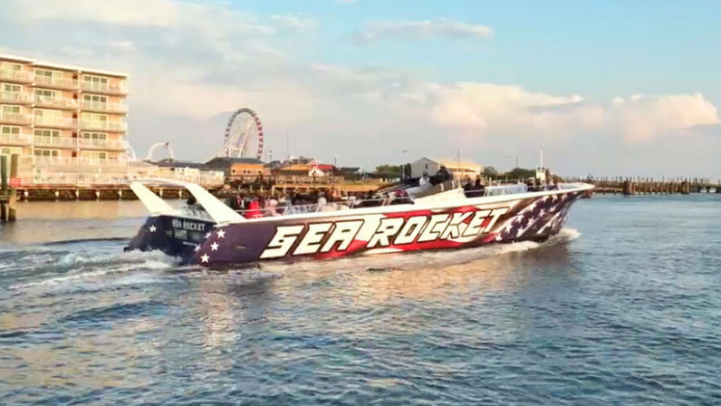 Patriotic red-white-blue speedboat with bold lettering cutting through water past a sunny seaside boardwalk and Ferris wheel at a waterfront pier