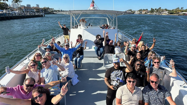 Group of smiling adults partying on an open-top boat cruising a sunny coastal waterway with palm-lined shore and waterfront homes, many raising drinks and hands under a clear blue sky with an American flag visible.
