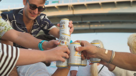 Friends cheering with white-and-gold beer cans on a sunny boat under a bridge — casual summer outdoor toast