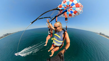 Two people parasailing over turquoise ocean and sandy coastline under a clear blue sky, colorful red-white-blue parachute, life vests and a speedboat wake visible below — aerial summer beach adventure.