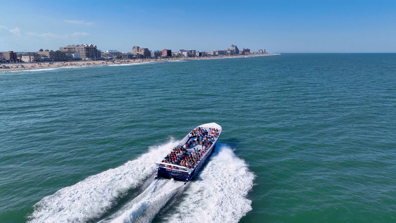 Crowded tour boat racing across blue-green ocean, leaving twin white wakes toward a busy sandy beach and beachfront skyline on a sunny day
