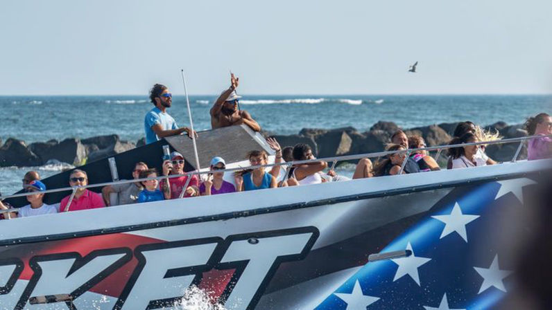 Crowded jet boat with star-spangled hull cruising along a rocky coast on a sunny day, passengers waving and enjoying the ocean ride with a seagull overhead.