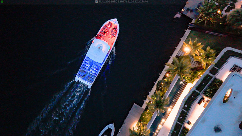 Aerial drone view of a lit tour boat cutting a foamy wake through dark water past a palm-lined waterfront marina at dusk, red and blue deck lights and string lights along the promenade