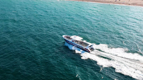 Aerial shot of a white-and-blue speedboat zipping across turquoise ocean, carving a foamy wake toward a sandy coastline.