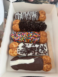 White bakery box holding five chocolate‑coated bubble‑waffle eclairs with toppings: white drizzle, crushed cookie crumbs, rainbow sprinkles, mini chocolate chips, and a white stripe — colorful dessert box.