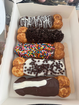 White bakery box holding five chocolate‑coated bubble‑waffle eclairs with toppings: white drizzle, crushed cookie crumbs, rainbow sprinkles, mini chocolate chips, and a white stripe — colorful dessert box.
