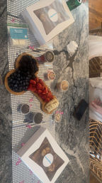 Playful dessert charcuterie on a marble kitchen countertop: wooden board with blueberries, blackberries, sliced strawberries and bananas, surrounded by small jars of sprinkles, nuts, chocolate chips and two boxed pastries.