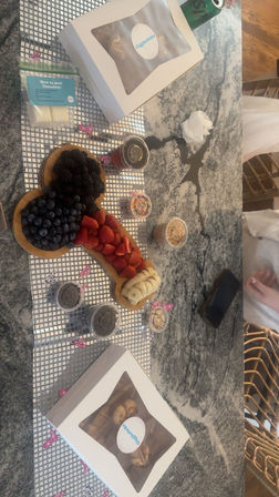 Playful dessert charcuterie on a marble kitchen countertop: wooden board with blueberries, blackberries, sliced strawberries and bananas, surrounded by small jars of sprinkles, nuts, chocolate chips and two boxed pastries.