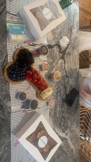 Playful dessert charcuterie on a marble kitchen countertop: wooden board with blueberries, blackberries, sliced strawberries and bananas, surrounded by small jars of sprinkles, nuts, chocolate chips and two boxed pastries.