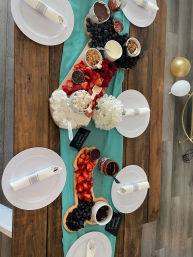Top-down view of a festive dessert grazing board on an indoor wooden table with a teal runner, piled with strawberries, blueberries, blackberries, raspberries, mini marshmallows, chocolate dipping sauces, granola and whipped cream, surrounded by white plates with napkins and gold and white balloons.