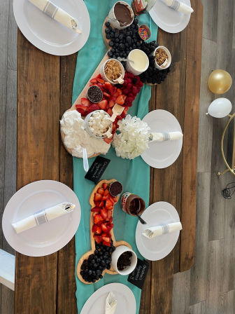 Top-down view of a festive dessert grazing board on an indoor wooden table with a teal runner, piled with strawberries, blueberries, blackberries, raspberries, mini marshmallows, chocolate dipping sauces, granola and whipped cream, surrounded by white plates with napkins and gold and white balloons.