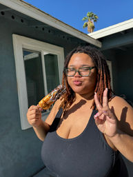 Person with glasses and brown dreadlocks in a black tank top holds a chocolate- and sprinkle-topped pastry on a stick, flashing a peace sign with a playful expression in a sunny backyard with a palm tree and blue sky.