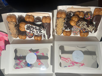 Two white pastry boxes filled with assorted bubble-waffle bites and chocolate-dipped eclairs topped with colorful sprinkles, cookie crumbs, slivered almonds and white icing drizzle — bright party dessert boxes on a table.