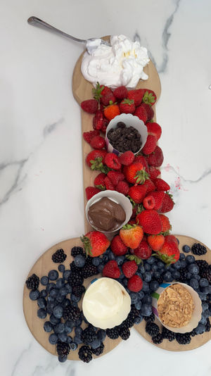 Playful wooden dessert charcuterie board on white marble countertop with fresh strawberries, raspberries, blueberries and blackberries, plus dollops of whipped cream, chocolate spread, yogurt and crunchy cookie crumbs.