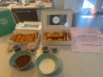 Two open boxes of golden pretzel sticks on a kitchen island with bowls of melted chocolate and white icing, small cups of sprinkles and toppings, and a pink confetti-patterned cloth nearby.
