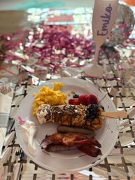 Festive brunch plate with scrambled eggs, bacon and sausage, chocolate‑drizzled waffle pop on a stick, fresh berries and whipped cream on a reflective confetti‑covered table with a pink flute