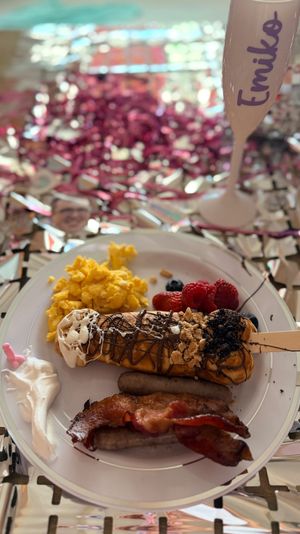 Festive brunch plate with scrambled eggs, bacon and sausage, chocolate‑drizzled waffle pop on a stick, fresh berries and whipped cream on a reflective confetti‑covered table with a pink flute