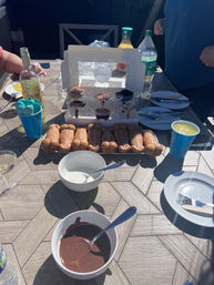 Sunny backyard dessert table with a fun fondue setup: row of novelty phallic-shaped pastries, bowls of melted chocolate and white dipping sauce, cups of toppings and bottles of drinks, plates and utensils ready for guests.