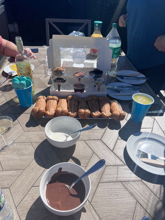 Sunny backyard dessert table with a fun fondue setup: row of novelty phallic-shaped pastries, bowls of melted chocolate and white dipping sauce, cups of toppings and bottles of drinks, plates and utensils ready for guests.