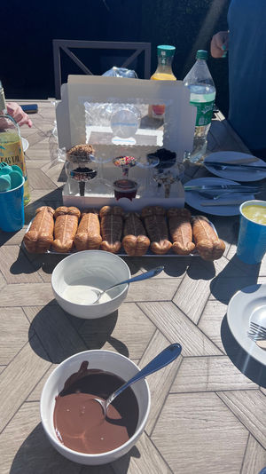 Sunny backyard dessert table with a fun fondue setup: row of novelty phallic-shaped pastries, bowls of melted chocolate and white dipping sauce, cups of toppings and bottles of drinks, plates and utensils ready for guests.