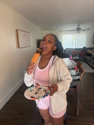 Person in pink activewear biting a chocolate-topped pastry on a stick, holding a crumb-filled paper plate in a bright open-plan living room with sofa and dining area behind.