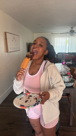 Person in pink activewear biting a chocolate-topped pastry on a stick, holding a crumb-filled paper plate in a bright open-plan living room with sofa and dining area behind.