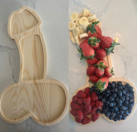 Playful penis-shaped wooden serving board on a marble countertop, arranged as a fruit platter with strawberries, banana slices, raspberries and blueberries.