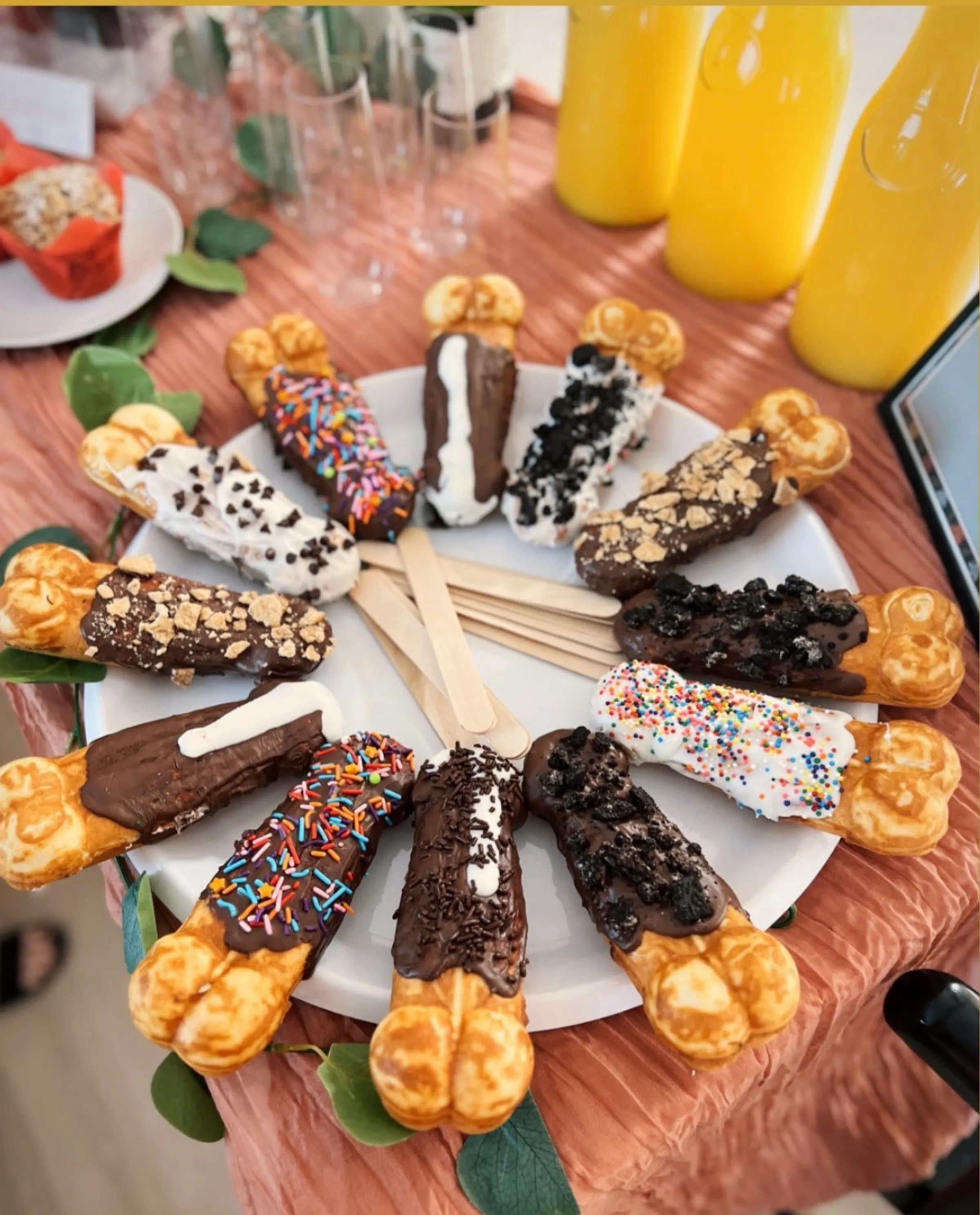 Colorful dessert display of assorted chocolate-dipped bubble waffle sticks on wooden sticks arranged on a white platter, topped with sprinkles, crushed cookies, chocolate chips and drizzles, with yellow juice bottles and a party table setting in the background.