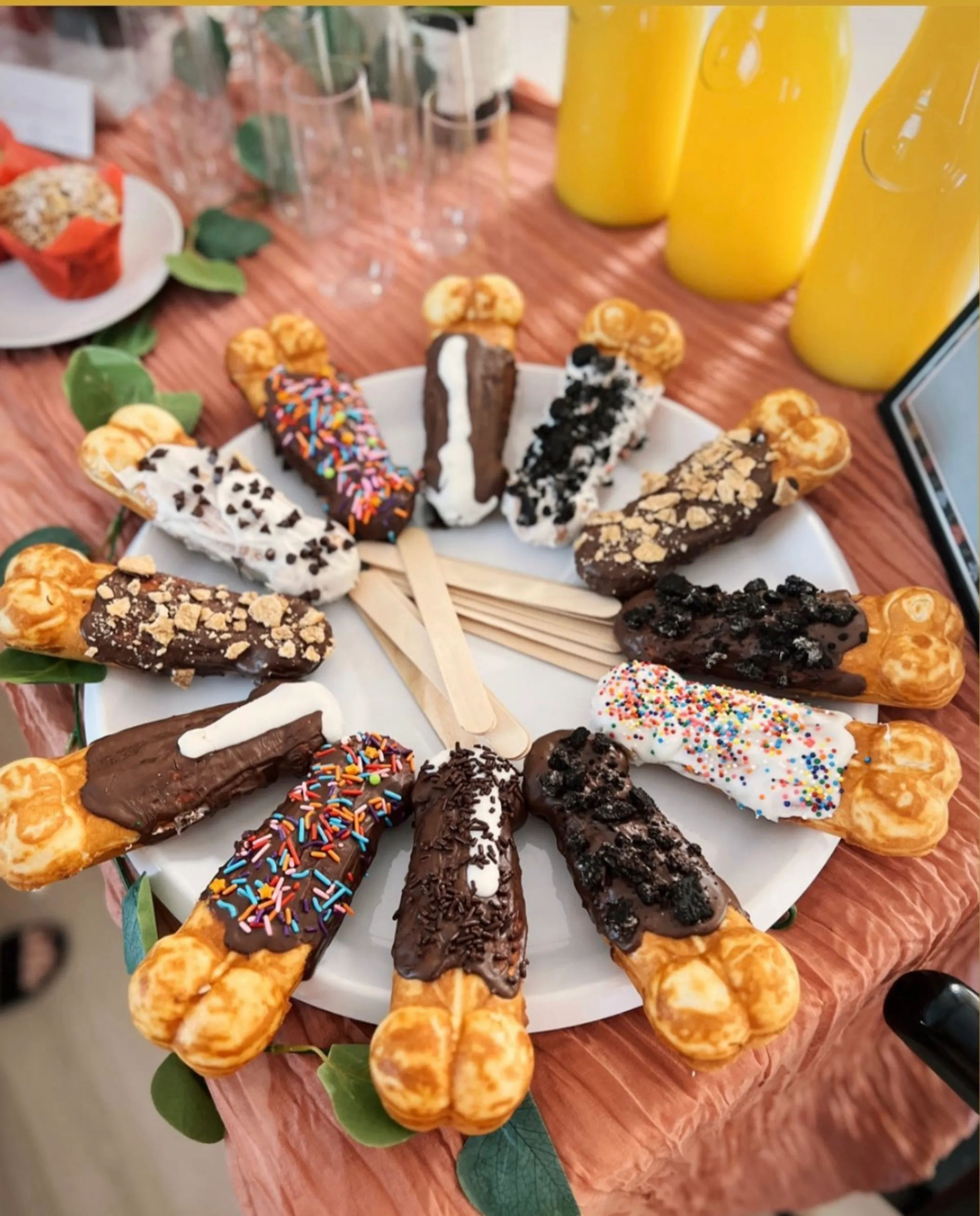 Colorful dessert display of assorted chocolate-dipped bubble waffle sticks on wooden sticks arranged on a white platter, topped with sprinkles, crushed cookies, chocolate chips and drizzles, with yellow juice bottles and a party table setting in the background.