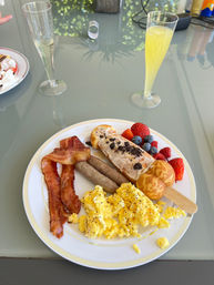 Brunch plate on a glass patio table: scrambled eggs, crispy bacon, breakfast sausages, fresh strawberries and blueberries, a cream-topped waffle pop with cookie crumbs, and two mimosas.