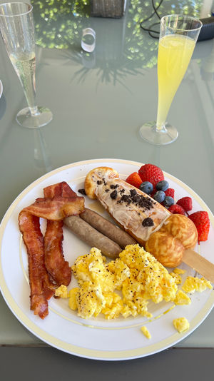 Brunch plate with fluffy scrambled eggs, crispy bacon, breakfast sausages, fresh strawberries, blueberries and raspberries, plus a cream-topped waffle pop on a white rimmed plate beside two champagne flutes with mimosa and sparkling wine on a glass table.