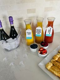 Mimosa brunch setup on a bright kitchen counter with champagne bottles on ice, labeled carafes of orange, passion‑orange & guava, and grapefruit juices, clear plastic flutes, bowls of blueberries, raspberries and sliced strawberries, and a plate of waffles.