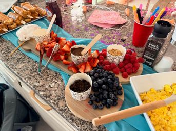 Colorful brunch spread on a granite kitchen counter: wooden boards piled with strawberries, raspberries and blueberries, paper cups of melted chocolate, granola and crushed cookies, mini pancake skewers, syrup bottle, scrambled eggs in a white dish and festive napkins.