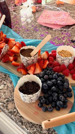 Colorful brunch spread on a granite kitchen counter: wooden boards piled with strawberries, raspberries and blueberries, paper cups of melted chocolate, granola and crushed cookies, mini pancake skewers, syrup bottle, scrambled eggs in a white dish and festive napkins.