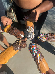 Overhead shot of chocolate-covered frozen bananas on cones with sprinkles, crushed cookies and cereal toppings, held together outdoors on a sunny sidewalk.