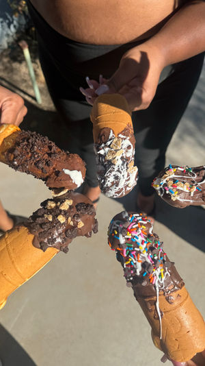 Overhead shot of chocolate-covered frozen bananas on cones with sprinkles, crushed cookies and cereal toppings, held together outdoors on a sunny sidewalk.