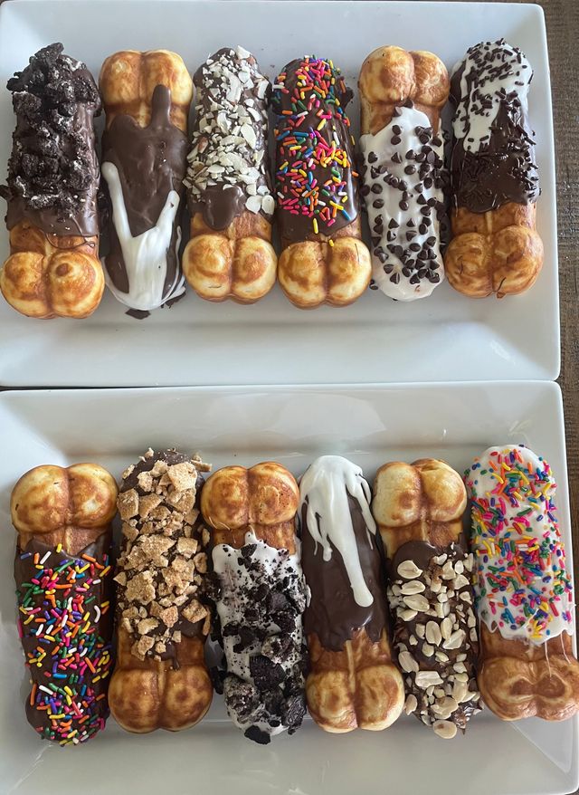 Close-up of two white plates with a colorful assortment of dipped bubble‑waffle sticks — chocolate and vanilla glazes topped with rainbow sprinkles, chopped nuts, crushed cookies and candies, gourmet dessert