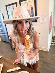 Playful woman in a pearl-trimmed pink hat holding a novelty toasted pastry on a stick in a bright home kitchen, posing for a party photo