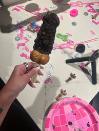Chocolate-covered ice cream pop with crushed cookie crumble and bubble-egg waffle base on a wooden stick, held over a party table strewn with pink confetti, toy figurines and a pink paper plate.