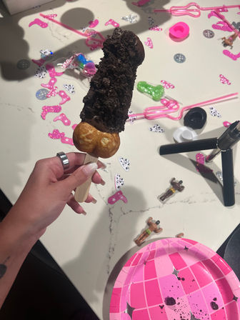 Chocolate-covered ice cream pop with crushed cookie crumble and bubble-egg waffle base on a wooden stick, held over a party table strewn with pink confetti, toy figurines and a pink paper plate.