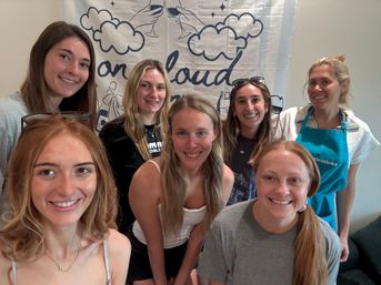 Cheerful group selfie of seven women indoors in front of a cloud-themed banner — casual gathering with smiling faces and bright natural light.