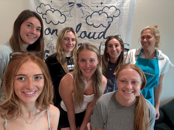 Cheerful group selfie of seven women indoors in front of a cloud-themed banner — casual gathering with smiling faces and bright natural light.