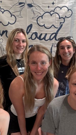 Cheerful group selfie of seven women indoors in front of a cloud-themed banner — casual gathering with smiling faces and bright natural light.