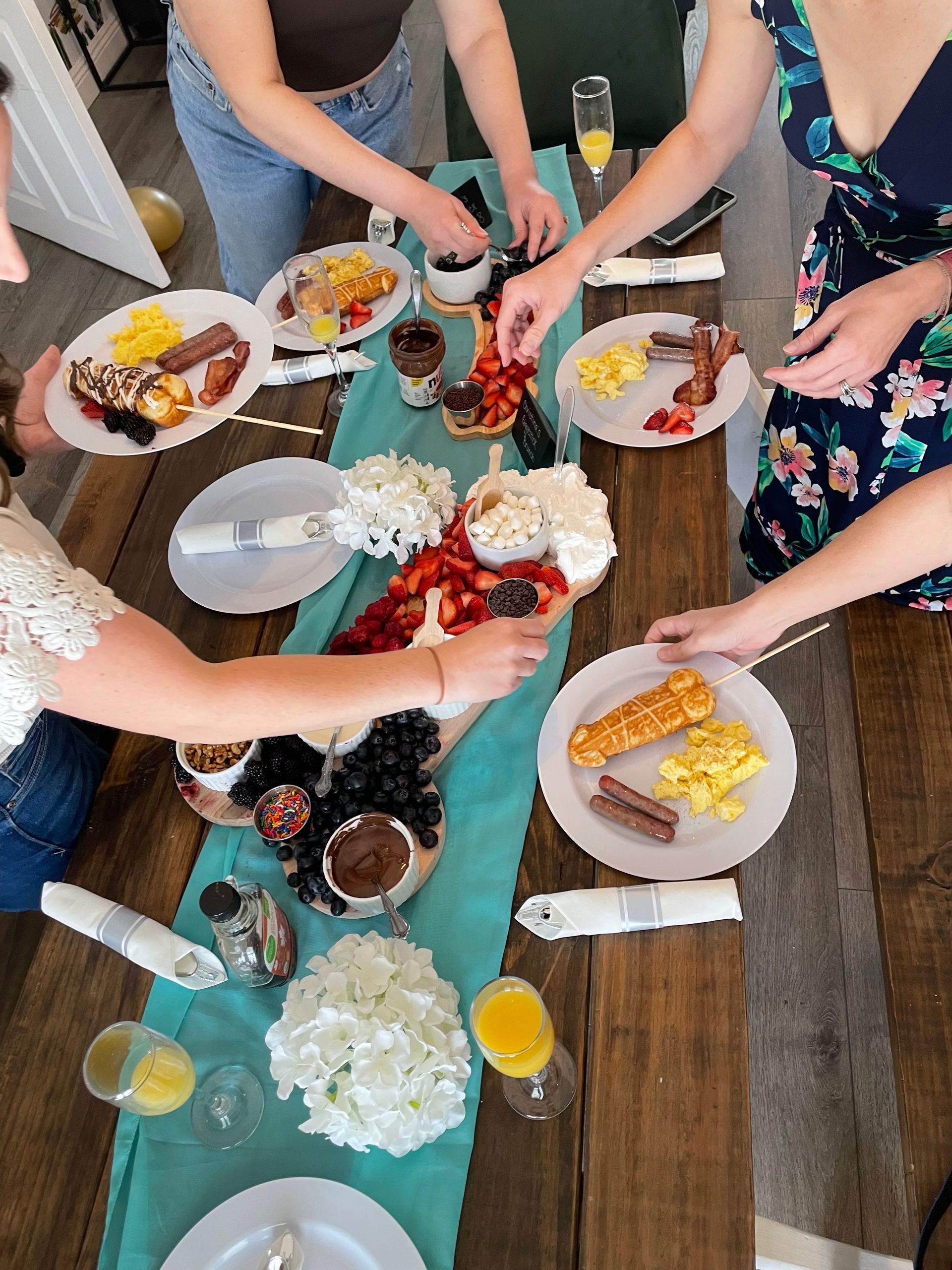 Top-down brunch scene on a wooden table with a teal runner — plates of scrambled eggs, sausages, bacon and waffle-on-a-stick, bowls of strawberries, blueberries, marshmallows, Nutella and chocolate sauce, sprinkles, white flower centerpieces and mimosas as hands reach to serve.