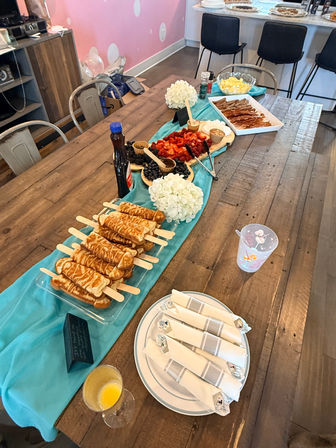 Indoor brunch buffet on a rustic wooden table with teal runner: pancake-on-a-stick stacks, fresh strawberries and blueberries, bowls of eggs, tray of bacon and sausages, syrup bottle, folded napkins and a mimosa glass — casual party setup.