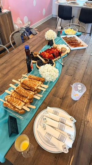Indoor brunch buffet on a rustic wooden table with teal runner: pancake-on-a-stick stacks, fresh strawberries and blueberries, bowls of eggs, tray of bacon and sausages, syrup bottle, folded napkins and a mimosa glass — casual party setup.