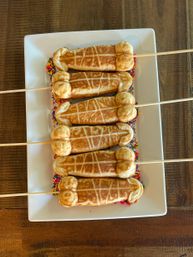 Playful row of six novelty phallic-shaped waffle pops on wooden skewers with rainbow sprinkles, arranged on a white rectangular plate on a rustic wooden table